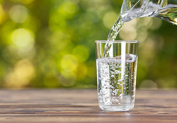 Water from jug pouring into glass on wooden table outdoors.