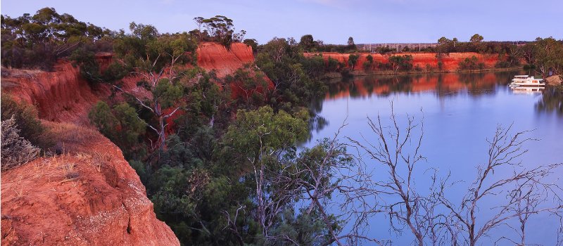 Red cliffs of Murray river on the border between Victoria and New South Wales states of Australia. Elevated red bank of the river overlooks river bend with cruise ship at sunset.