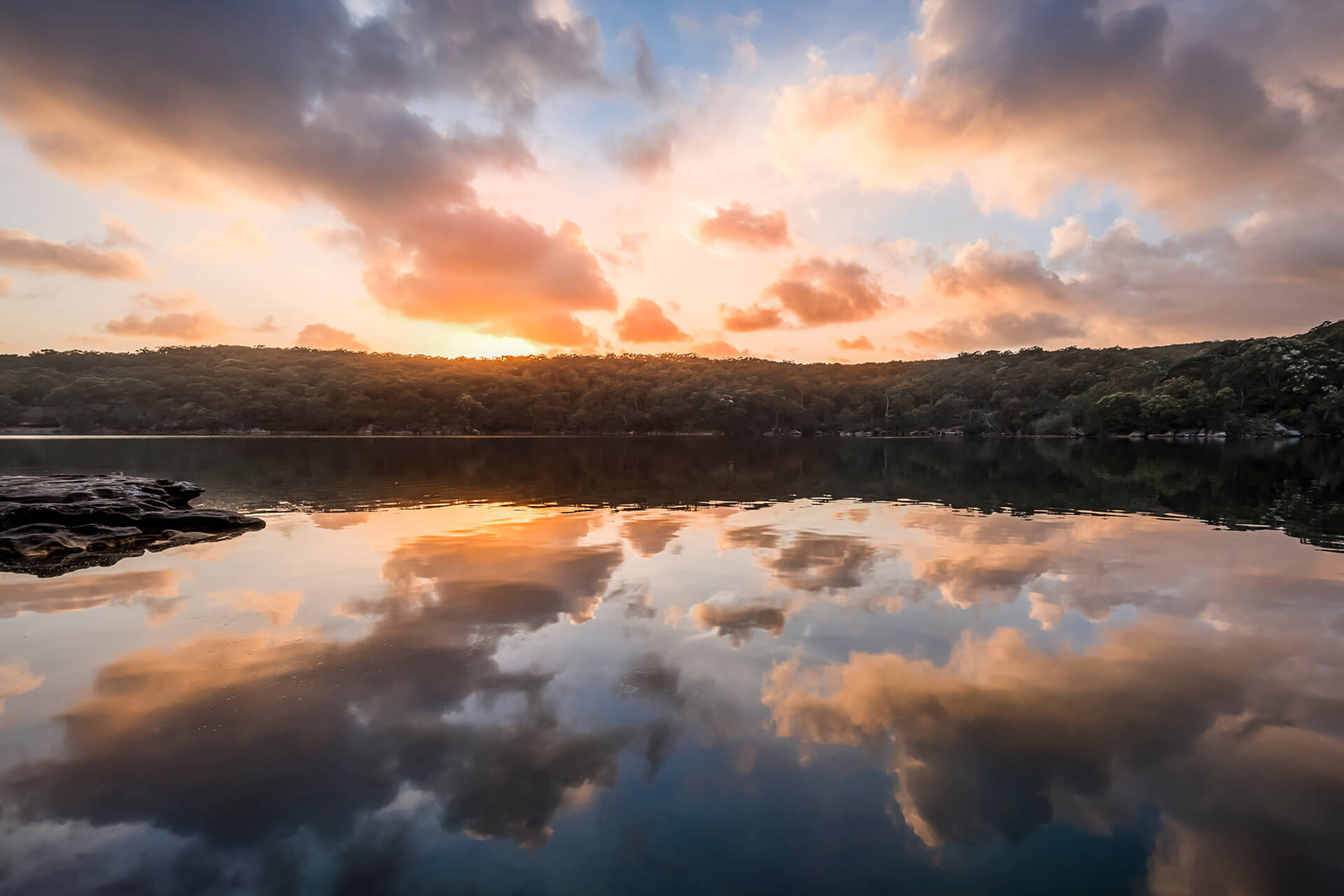 A lake and hill bank with an orange sunset. The clouds are reflected in the water
