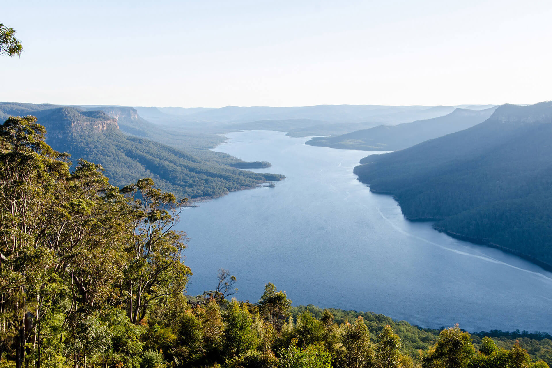 a winding lake of blue water surrounded by tall hills that are covered in trees. The sky is little gray. Lake Burragorang, NSW.