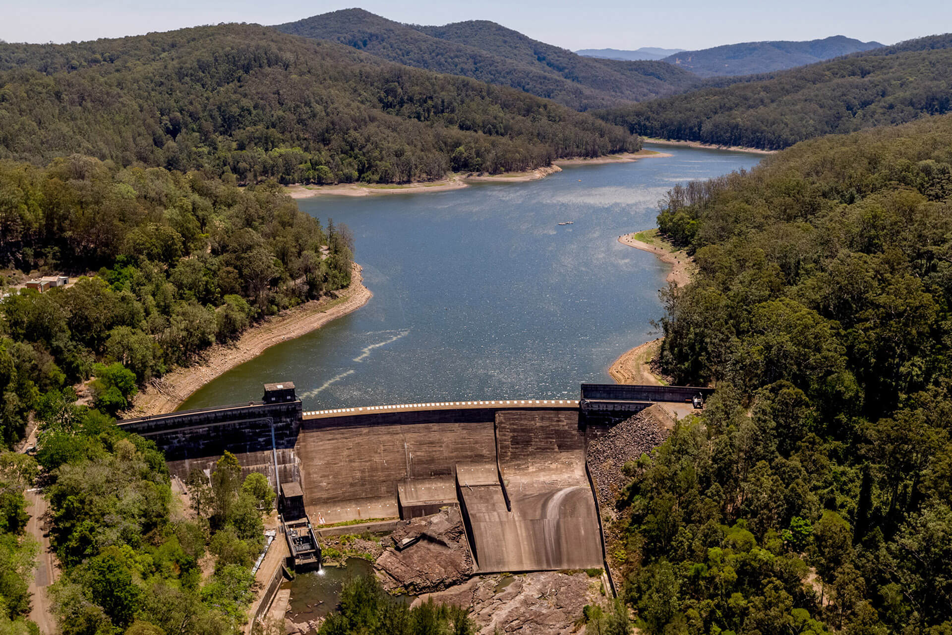 Body of water behind a large concrete dam wall. The water is surrounded by tree-covered hills