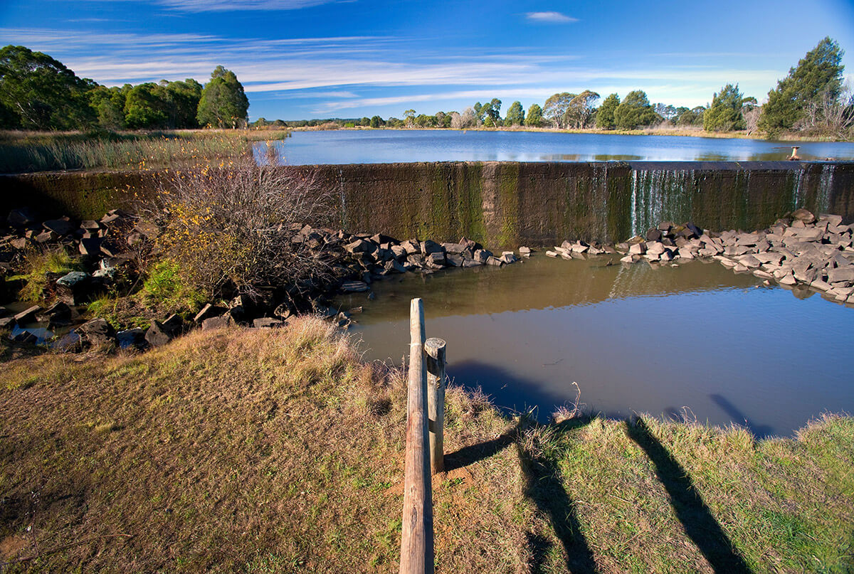 A concrete weir holds back water with blue sky and trees in the backrgound