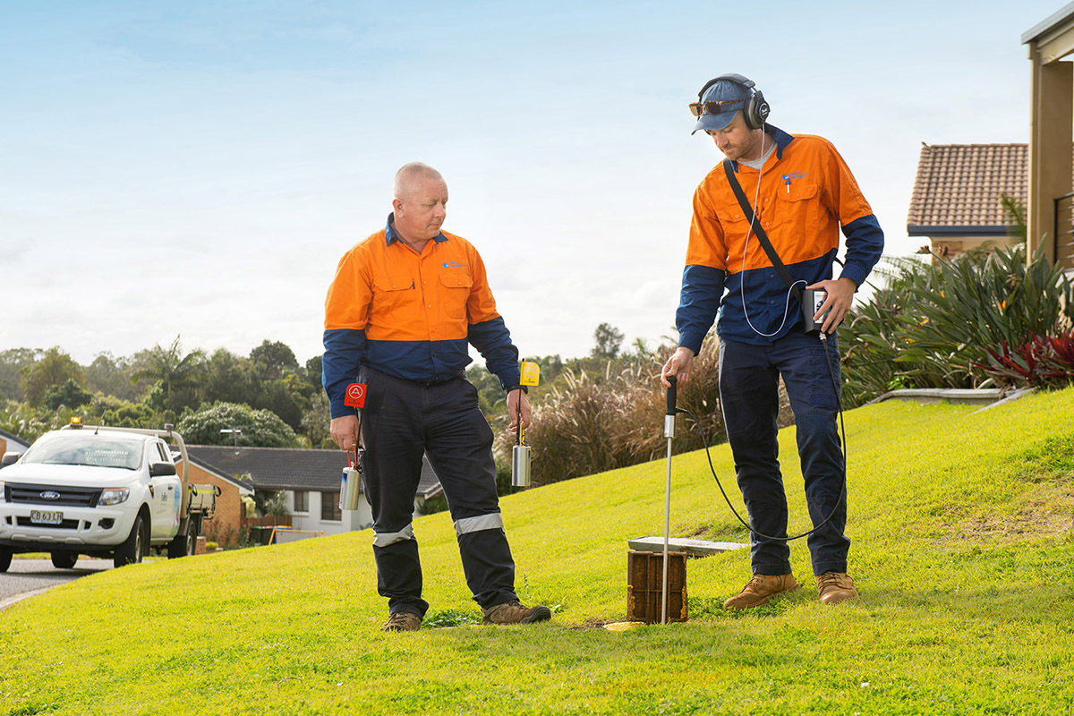 Two men in orange shirts use equipment to look for leaks