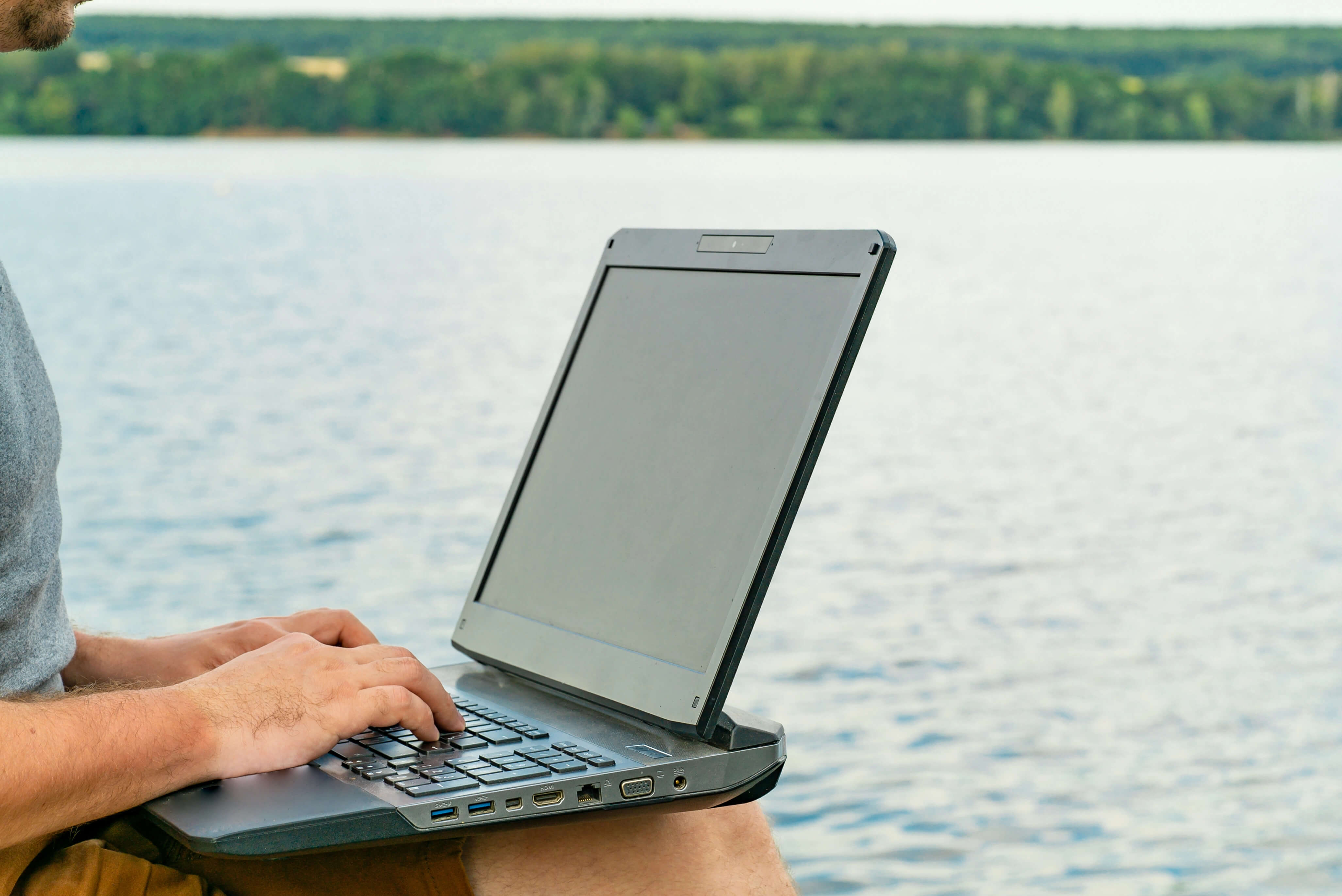 a person sits nexts to water typing on a laptop sitting on their lap