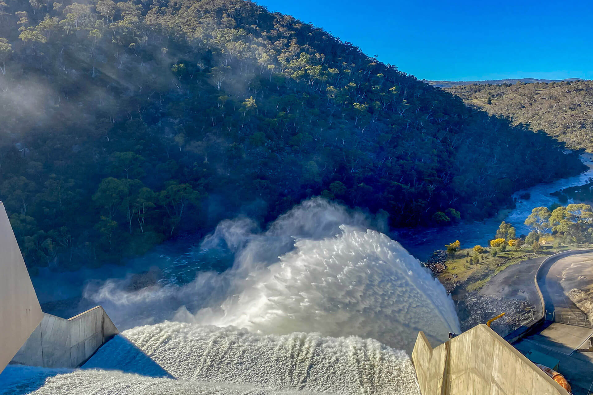 In the foreground water is being sprayed into the air in a fan shape. In the background is a hill covered in trees. The sky is clear and blue. This is a photo from the Snowy Mountains.