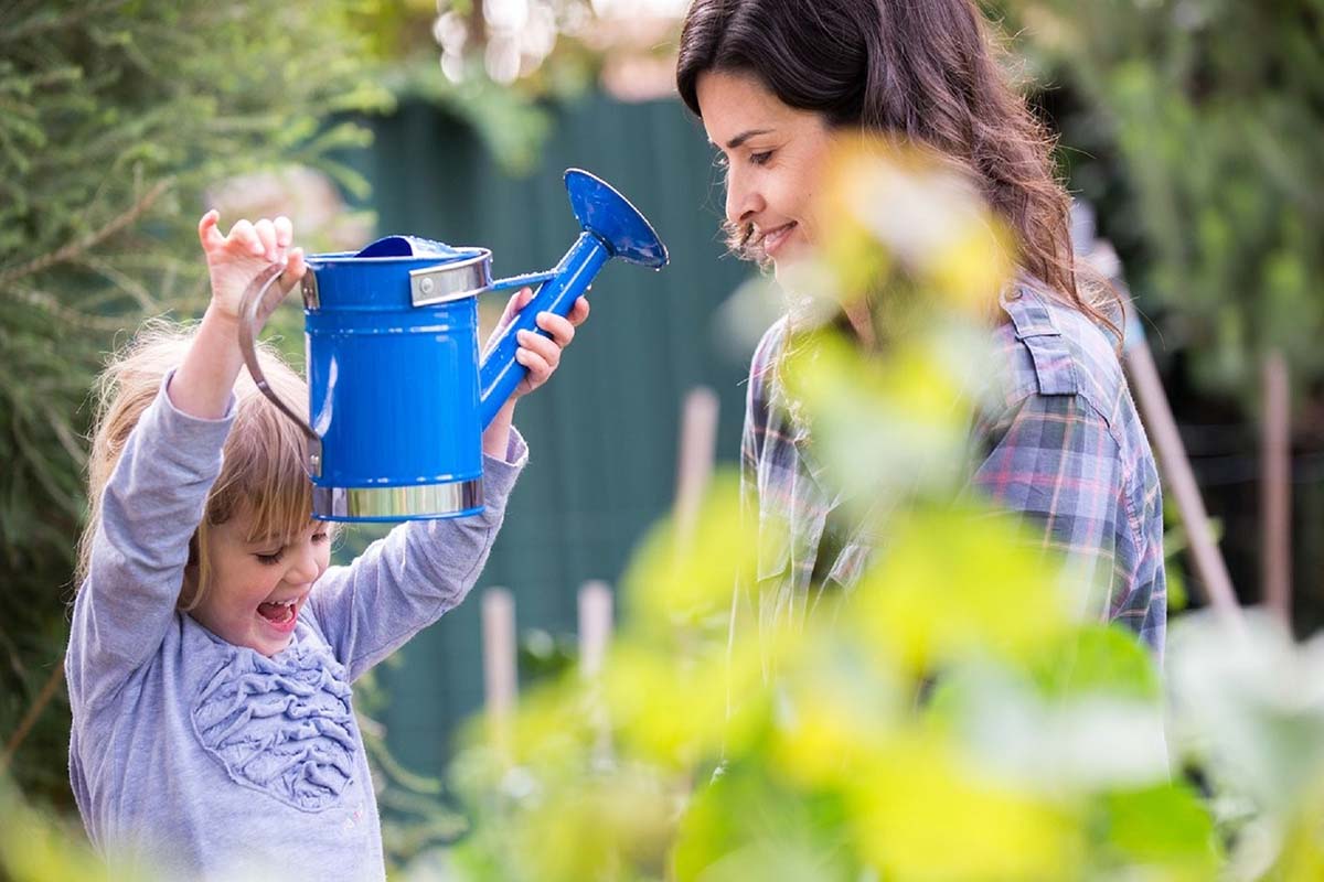 a girl in a purple shirt holds a blue watering can next to an adult woman. Both a smiling