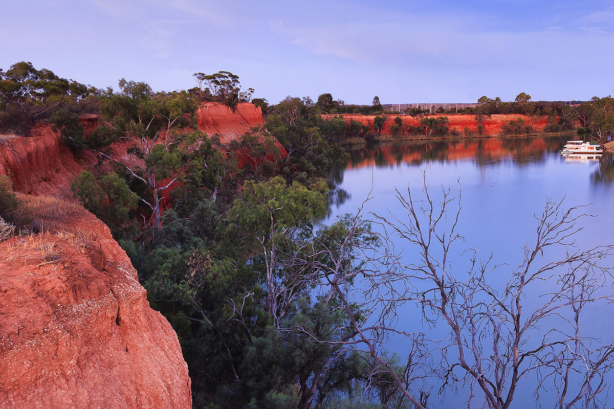 Red cliffs of Murray river on the border between Victoria and New South Wales states of Australia. Elevated red bank of the river overlooks river bend with cruise ship at sunset.